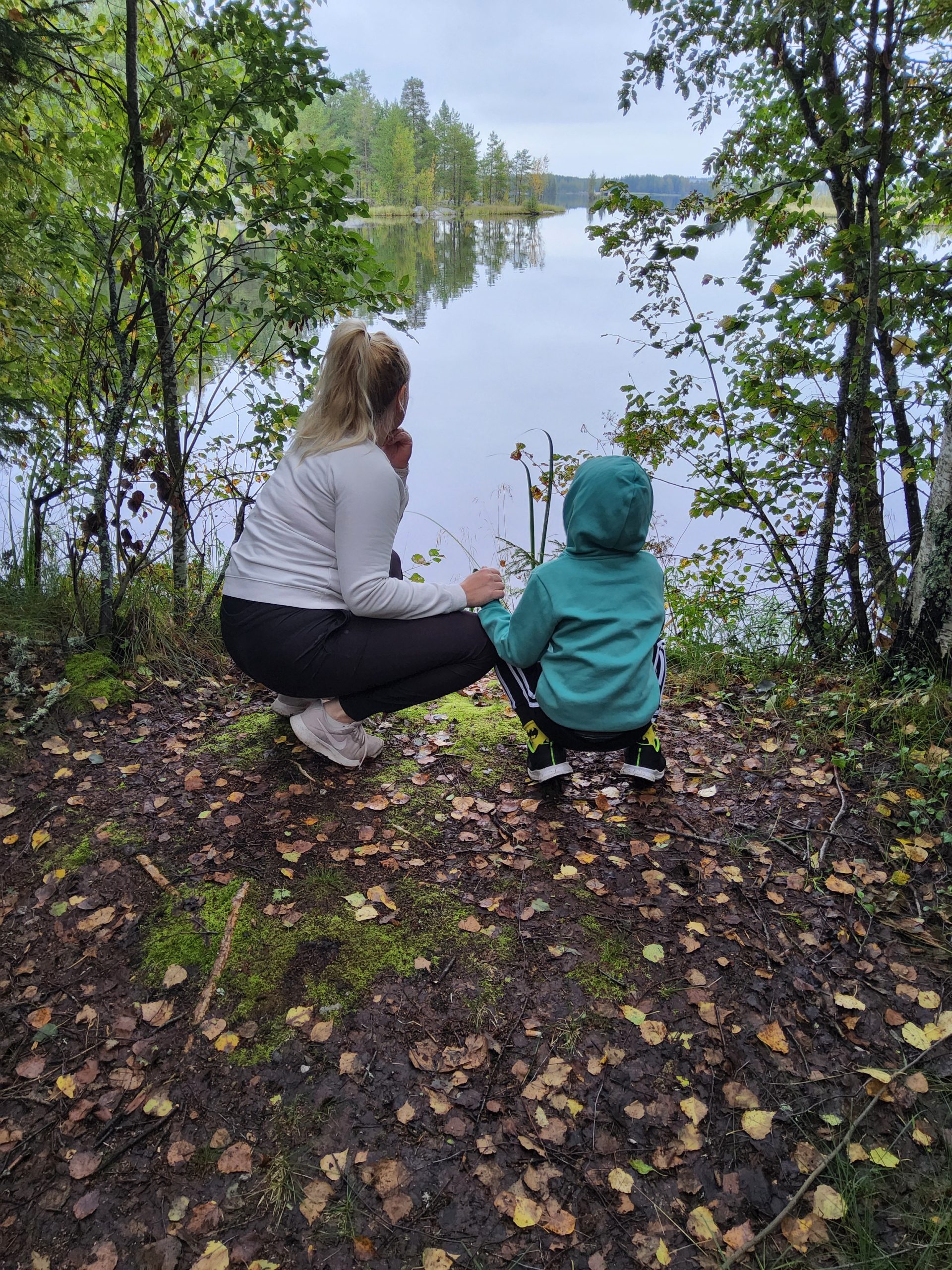 Nainen ja lapsi kyykyssä järven rannalla katselemassa järven maisemaa