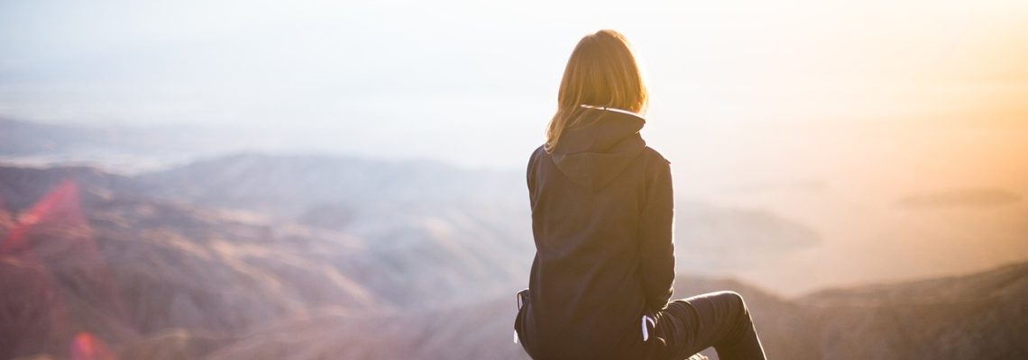Nainen istuu vuorenhuipulla ja katsoo kaukaisuuteen